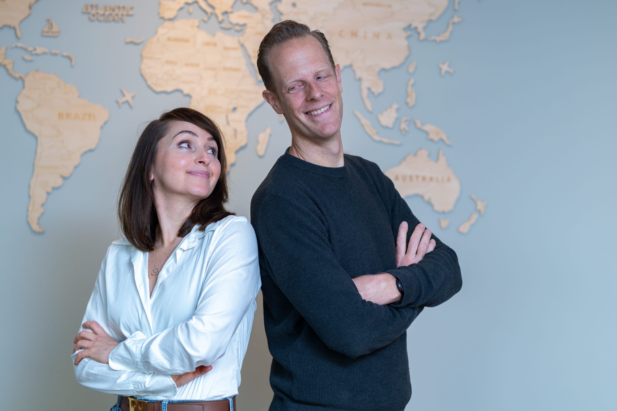 Dr. Kathrin und Dr. Claus Mangelsdorf, das Inhaber-Duo der Praxis, Rücken an Rücken vor einer Weltkarte aus Holz